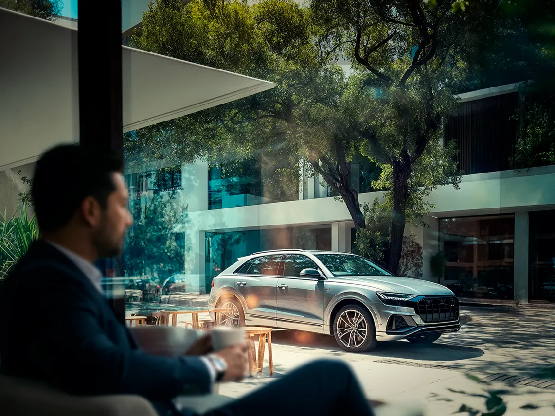 Hombre relajado tomando café en una terraza con su camioneta SUV estacionada al fondo, transmitiendo tranquilidad.